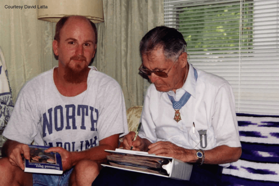 Color photo of Desmond Doss seated indoors, wearing a white shirt and his Medal of Honor around his neck, signing a binder with a pen. Next to him sits a man in a "North Carolina" T-shirt holding a book titled Desmond Doss: Conscientious Objector. A caption in the top left reads, "Courtesy David Latta."