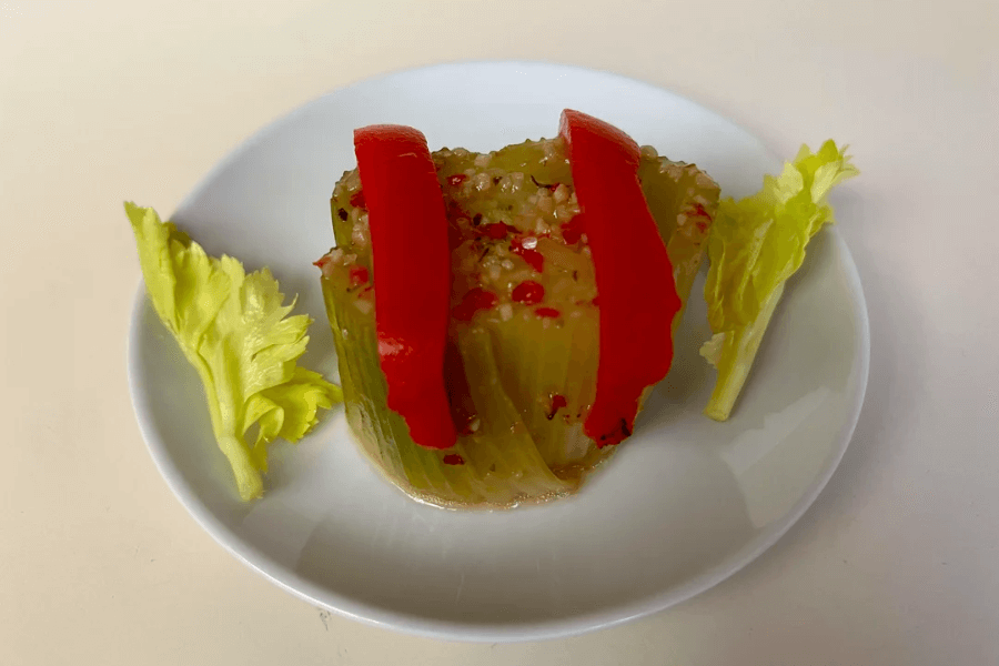 A molded savory gelatin made with chopped celery, pimentos, and seasoning, topped with two red pepper strips. The gelatin is served on a small white plate with two fresh celery leaves as garnish.