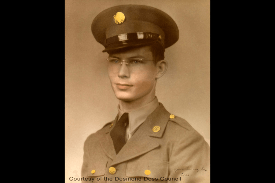 Sepia-toned portrait of a young man in a U.S. Army uniform, wearing a peaked cap with an eagle insignia, round glasses, and a tan dress coat with brass buttons and unit patches. He looks composed and proud, with the caption “Courtesy of the Desmond Doss Council” at the bottom and a faint handwritten note on the right side.