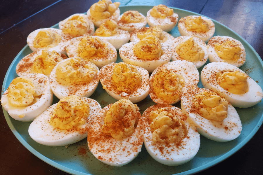 A teal plate filled with deviled eggs, each halved hard-boiled egg topped with a creamy yellow yolk mixture and a sprinkle of paprika. The eggs are neatly arranged in circular layers on a dark wooden table.