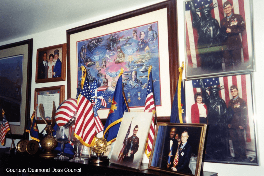 A colorful indoor display featuring numerous framed photos, patriotic memorabilia, and miniature flags, including American and state flags. Prominently featured are portraits of Desmond Doss in uniform, including ones taken beside a Statue of Liberty backdrop, as well as a tribute collage with military scenes and figures. A small placard notes, "Courtesy Desmond Doss Council."