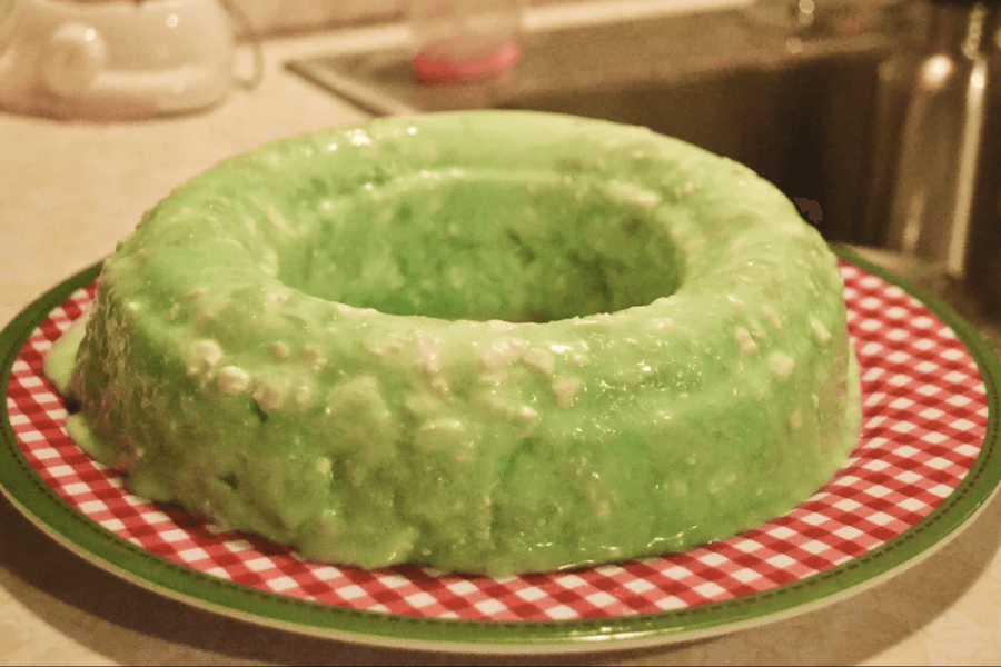 A retro-style green gelatin mold shaped into a ring and served on a red and white checkered plate. The Jell-O is flecked with visible bits of what appears to be cottage cheese or crushed pineapple, giving it a lumpy texture.