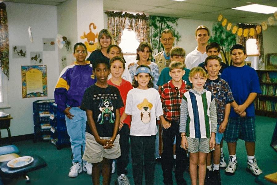 A group of schoolchildren pose for a photo inside a colorful, decorated classroom, with a few adults and Desmond Doss—wearing a tan military uniform and medal—standing at the back. The room is filled with learning posters, bookshelves, and hanging student artwork, creating a warm and educational environment.