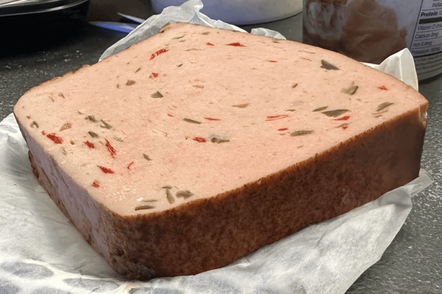A thick, rectangular block of olive loaf luncheon meat, speckled with green olives and red pimentos, resting on wax paper atop a kitchen counter. The meat has a smooth, pale pink texture with a darker crust along the edges.