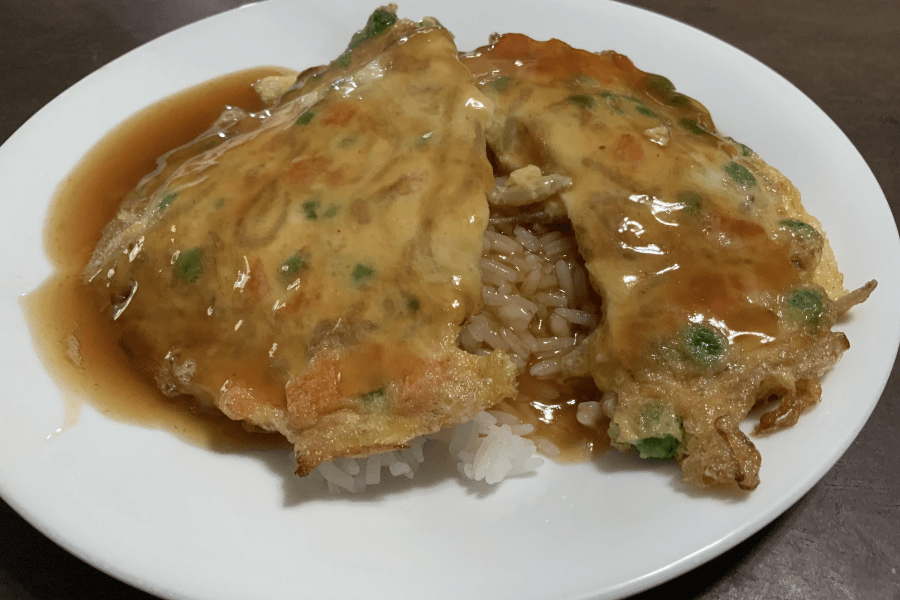 A plate of Japanese tenshindon featuring a fluffy omelet mixed with peas and carrots, draped over a bed of white rice and covered in a glossy, savory brown sauce.