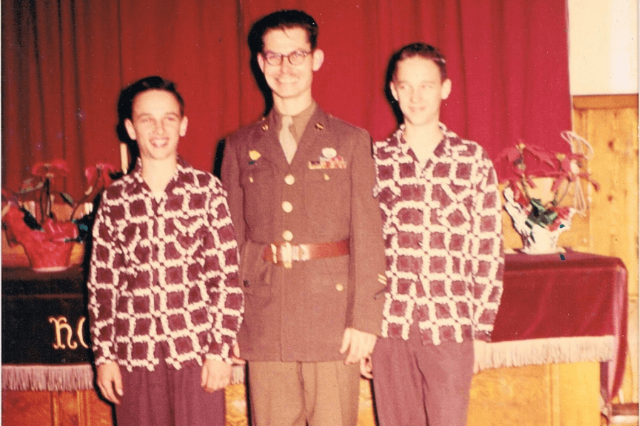 Desmond Doss stands smiling in full military uniform between two teenage boys wearing matching patterned shirts and dark trousers. The group is indoors in front of red curtains and decorative plants, likely during a church or community event.