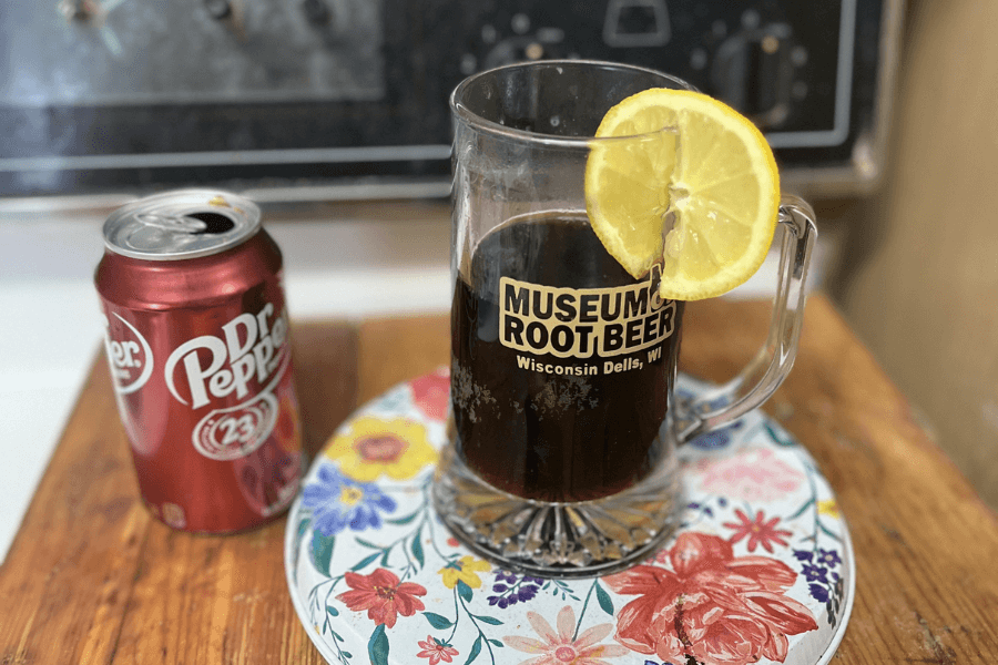 A can of Dr. Pepper next to a glass mug from the Museum of Root Beer in Wisconsin Dells, WI, filled with dark soda and garnished with a lemon slice, all placed on a colorful floral coaster on a wooden surface.