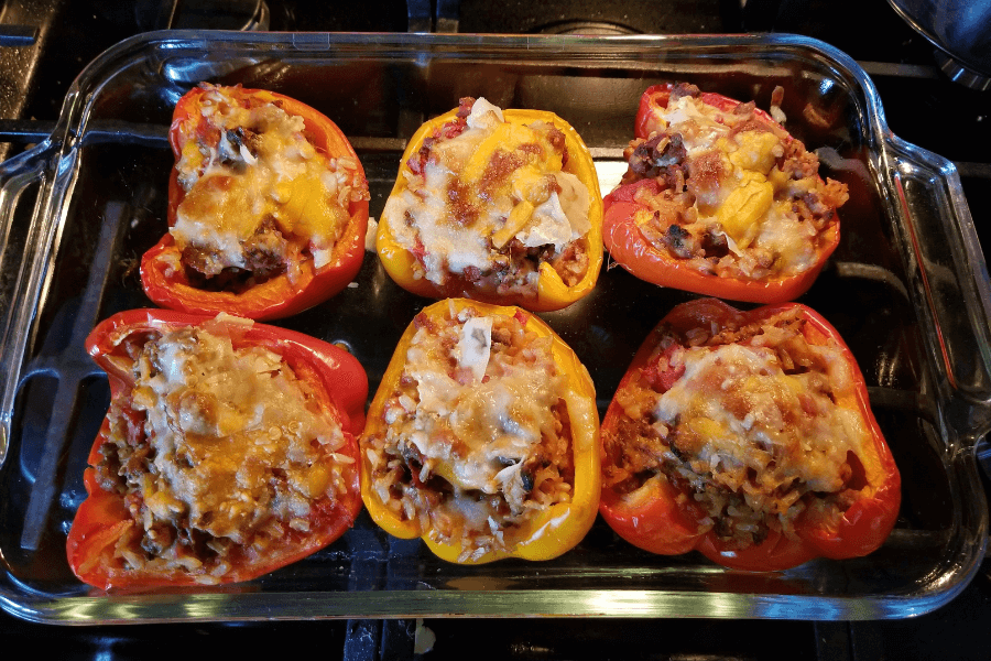 A glass baking dish filled with six halved bell peppers in red and yellow, stuffed with a hearty mixture of ground meat, rice, diced vegetables, and topped with melted cheese, fresh out of the oven.