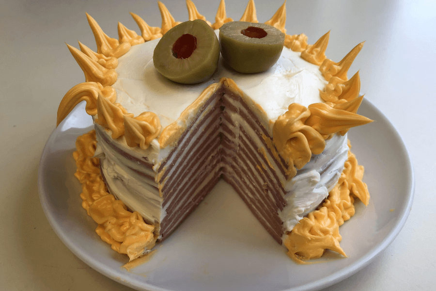 A layered savory cake made entirely of stacked slices of ham, frosted with white cream cheese or mayo, and decorated with piped orange cheese spread and two green olives on top, resembling a quirky meat-based celebration cake.