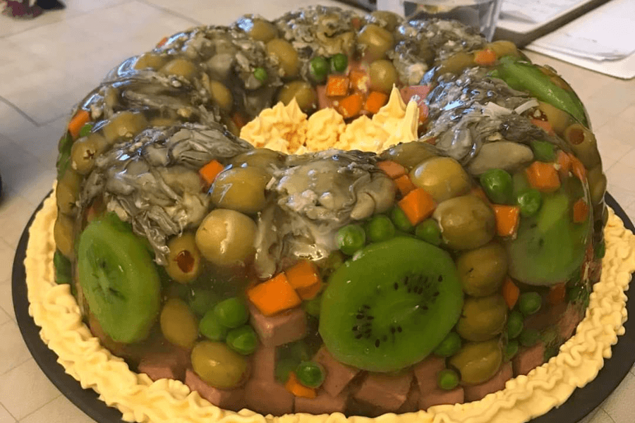 A vintage-style savory gelatin mold in a bundt shape, filled with an unusual combination of ingredients including green olives, canned oysters, peas, diced ham, carrot cubes, and slices of kiwi, all suspended in a clear gelatin. The base of the mold is piped with a yellow cream or mayonnaise-like garnish.