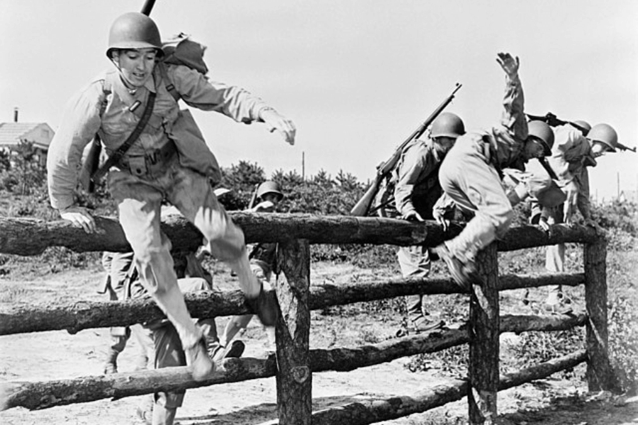 Black-and-white photo of U.S. soldiers in full gear leaping over a wooden obstacle during a World War II-era military training exercise. Each soldier wears a helmet and backpack, with some holding rifles as they vault over the fence in mid-air action. The scene captures a moment of intense physical training on a sunny day.