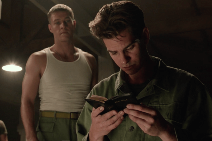 Scene from a military barracks where a soldier in green fatigues intently reads a small Holy Bible, while another man in a white tank top and army pants stands behind him, watching. The dimly lit room creates a solemn, focused atmosphere, highlighting the significance of the moment.