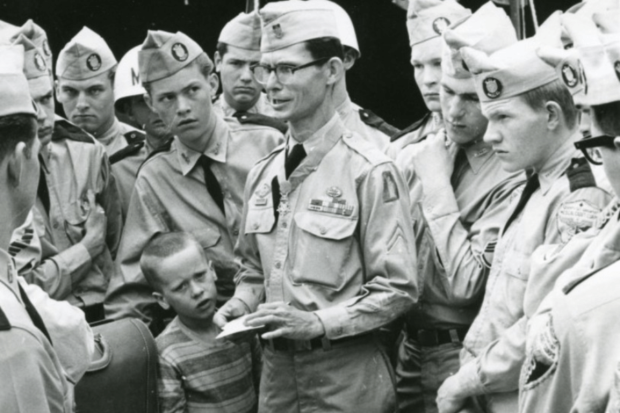Black-and-white photo of a decorated U.S. Army soldier in uniform speaking to a group of younger soldiers and cadets, with a young boy standing closely by his side. The group appears attentive, gathered in a semi-circle around the central figure, who wears several service ribbons and a Combat Medic badge.