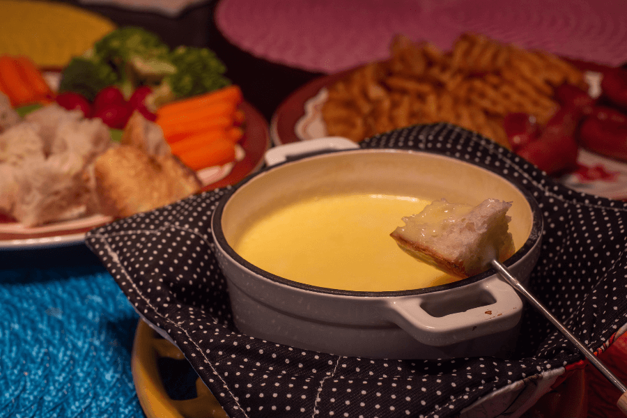 A pot of melted cheese fondue with a piece of bread on a skewer being dipped, placed in a white ceramic bowl wrapped in a black polka-dot cloth. Surrounding plates hold dippable items including waffle fries, carrots, broccoli, radishes, sausage, and chunks of bread.