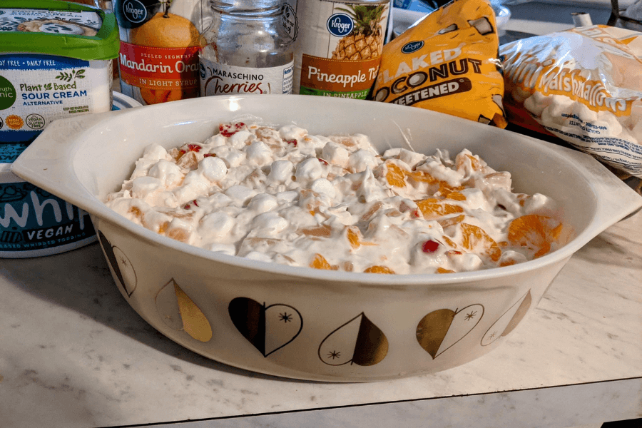 A vintage Pyrex bowl filled with ambrosia salad, a creamy mixture of whipped topping, mini marshmallows, mandarin oranges, maraschino cherries, pineapple tidbits, and shredded coconut. Behind the bowl are the ingredient containers, including whipped topping, vegan sour cream, and bags of marshmallows and coconut.