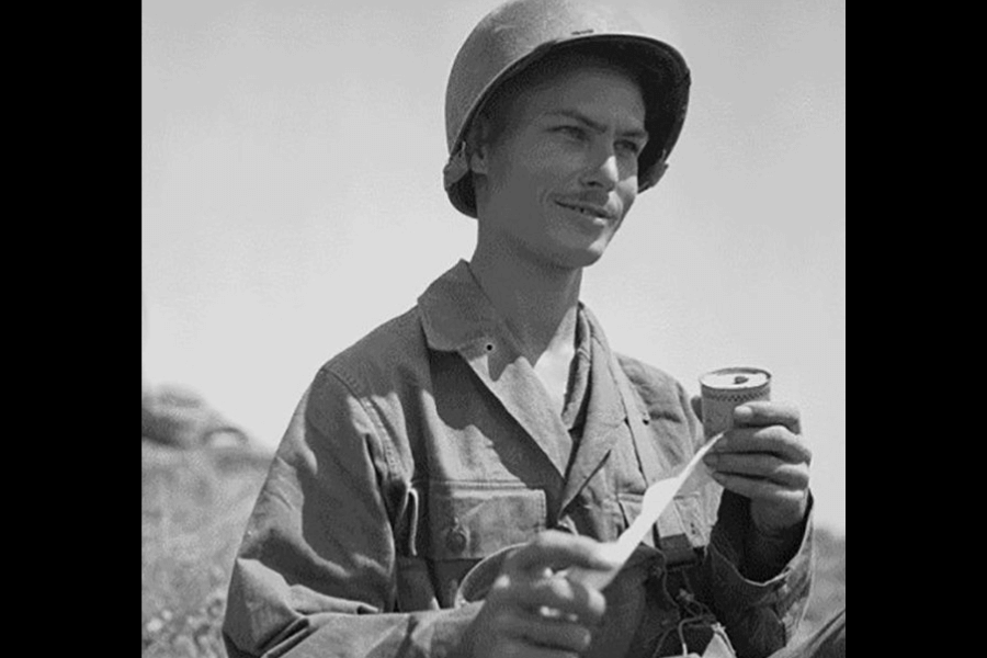 Black-and-white photo of a young U.S. Army soldier wearing a helmet and uniform, smiling while holding a roll of medical tape and pulling a strip from it. The setting appears to be outdoors during daylight, possibly in a wartime environment.