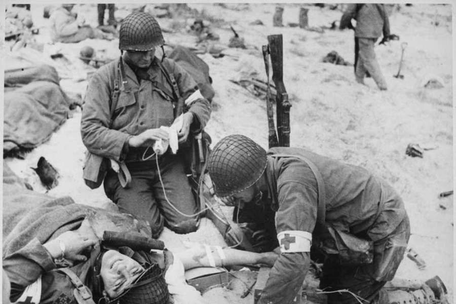 Black-and-white WWII photo showing two Army medics tending to a wounded soldier lying on the sandy ground. One medic is administering an IV drip while the other checks the soldier’s condition; both wear Red Cross armbands and combat gear. The chaotic battlefield is littered with gear and other wounded soldiers in the background.