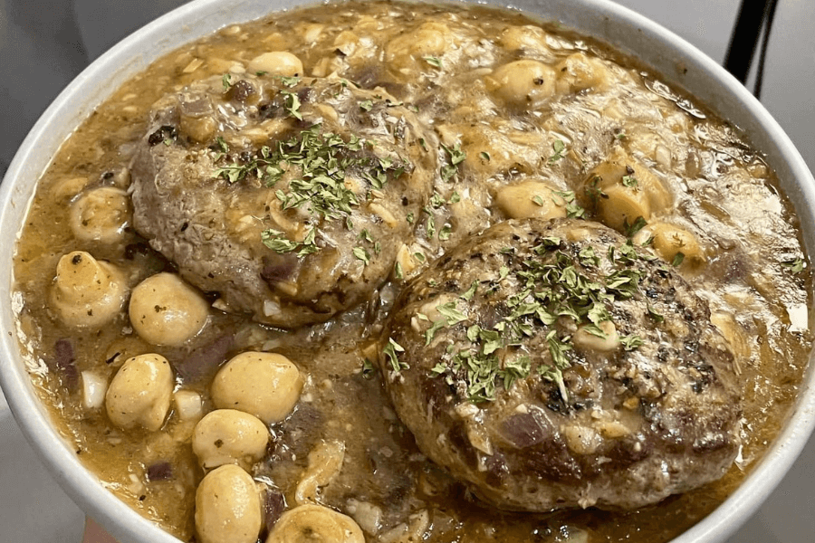 A bowl of hearty stew featuring two seared beef patties topped with dried herbs, surrounded by chickpeas, diced onions, and a thick, savory brown gravy. The dish appears rich and comforting, with visible seasoning and texture.