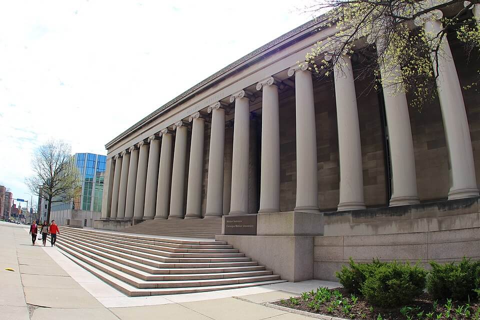 A grand neoclassical building with tall white columns lines a wide sidewalk, with three people walking past. A sign near the stairs reads “Walker Memorial, Campus Media University,” and a modern glass building stands in the background.