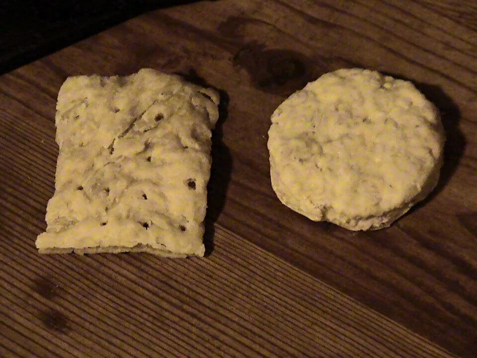 Two rustic, pale-colored oatcakes placed on a wooden surface. One oatcake is rectangular with visible holes, while the other is round and slightly thicker.