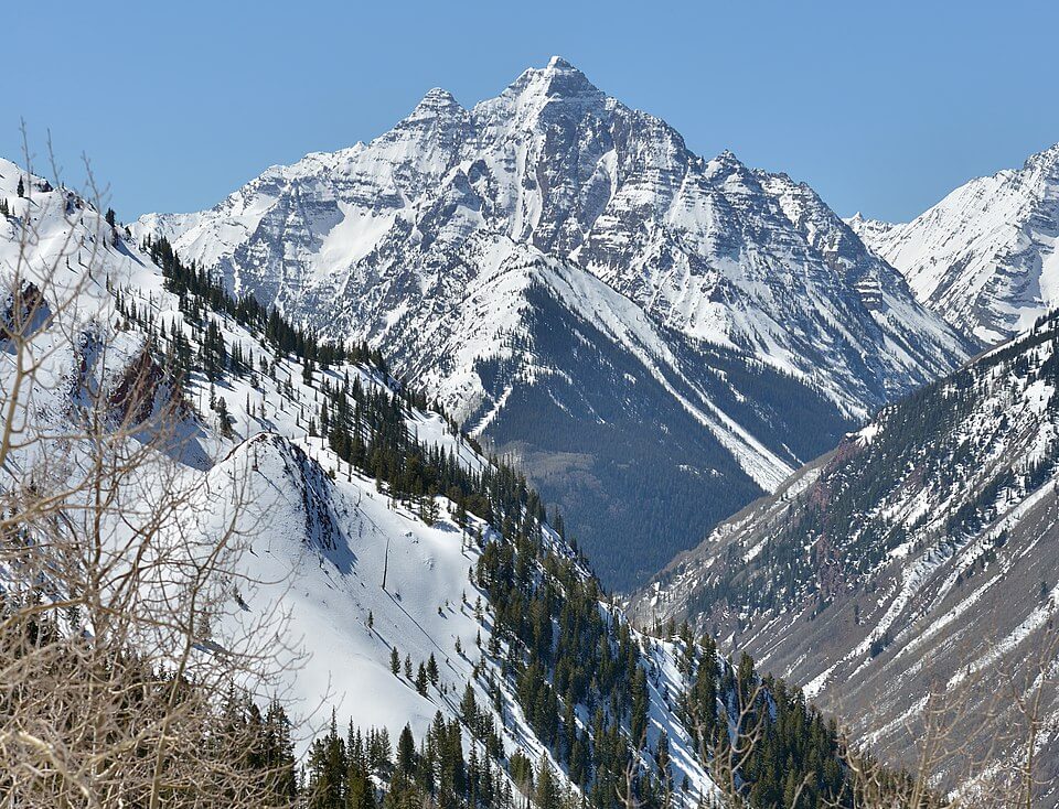 A towering, snow-covered mountain peak rises sharply against a clear blue sky. The surrounding slopes are blanketed in snow with dense pine forests and a few bare trees in the foreground.