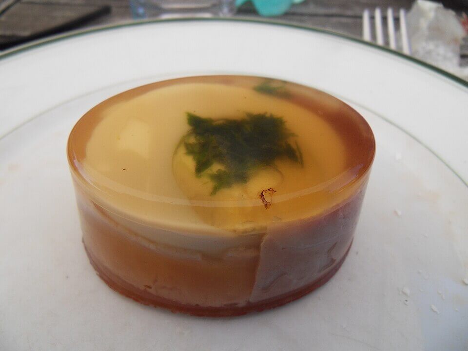 A circular, translucent aspic with a garnish of green herbs suspended inside. The jelly has a glossy surface and layers beneath, resting on a white plate with a green rim.