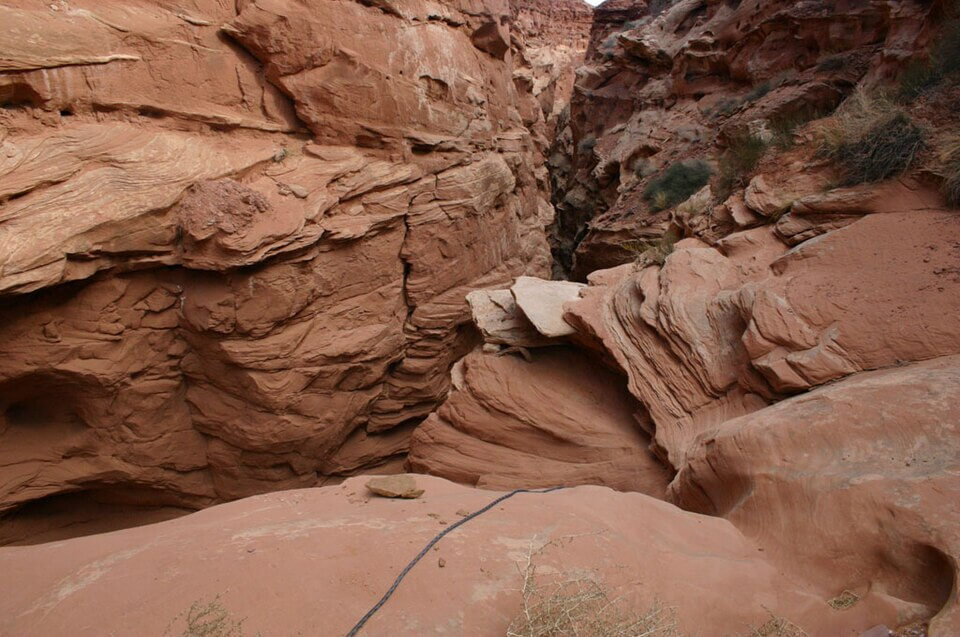 A rugged, narrow desert canyon with steep red rock walls and swirling sandstone formations. A climbing rope lies on the ground, hinting at the challenging terrain ahead.