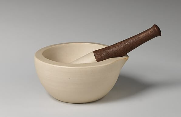 A beige ceramic mortar with a matching pestle featuring a dark wooden handle rests on a neutral gray background. The mortar has a small spout on one side and a smooth, matte finish.