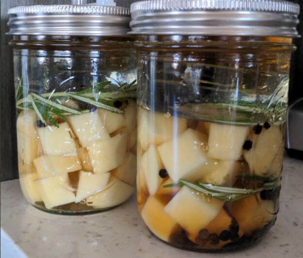 Two glass jars filled with cubed cheese submerged in oil sit on a countertop. The jars contain sprigs of rosemary and whole peppercorns, sealed tightly with metal lids.