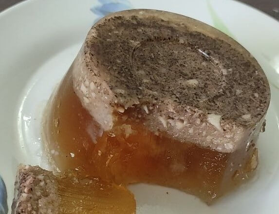 A round serving of meat aspic sits on a white plate with floral decoration. The gelatinous dish has a clear amber base and a darker, meaty layer on top, with one slice cut out and placed beside it.