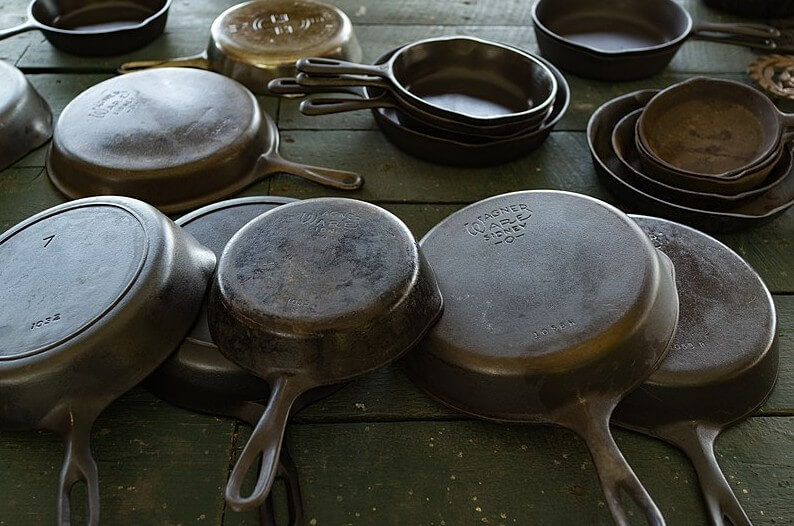 A collection of vintage cast iron skillets of various sizes lies on a rustic green wooden table. Some pans are stacked, while others are flipped to show embossed brand names like "Wagner Ware" and model numbers on their bases.