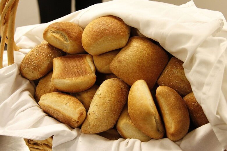 A woven basket lined with a white cloth is filled with a variety of golden-brown bread rolls. The rolls are slightly different in shape and texture, some with smooth tops and others dusted with flour or seeds.