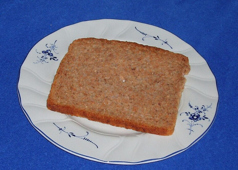 A single slice of brown bread placed on a white plate with blue floral designs. The bread appears plain and unbuttered, with a rough texture and one corner slightly bitten off.