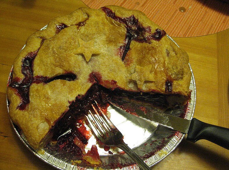 A partially sliced berry pie in a disposable aluminum tray, with dark red filling oozing out of the golden-brown crust. A fork and a knife rest beside the missing slice, with berry juice smeared on the tray.