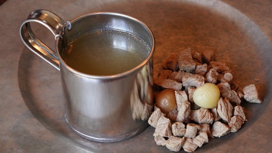 A metal cup filled with clear broth sits on a rustic metal plate. Next to the cup is a small pile of diced cooked meat and two whole pearl onions.