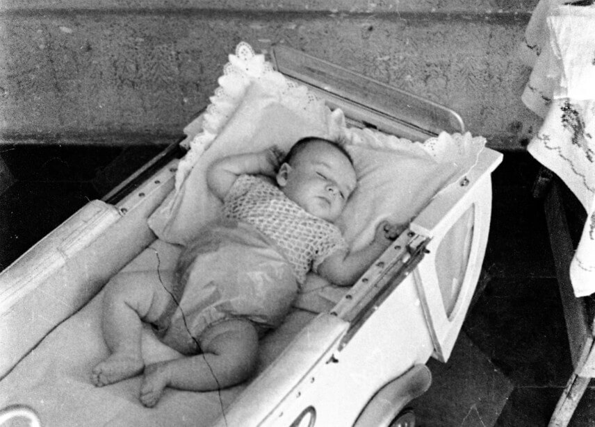 A black-and-white photo of a baby peacefully sleeping in a vintage pram, wearing a crocheted top and diaper cover, with lace-trimmed bedding surrounding them.







