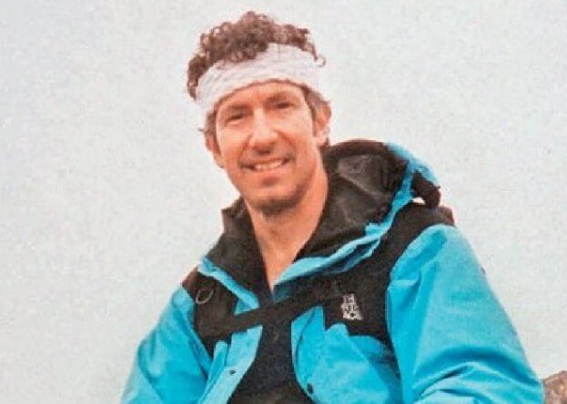  A smiling man with curly hair wears a light blue and black mountaineering jacket and a white headband, sitting outdoors with a cloudy sky in the background.
