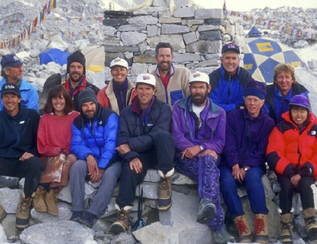  A group of mountaineers dressed in colorful cold-weather gear pose for a photo at a rocky Everest Base Camp, with tents, prayer flags, and snow-covered terrain in the background.