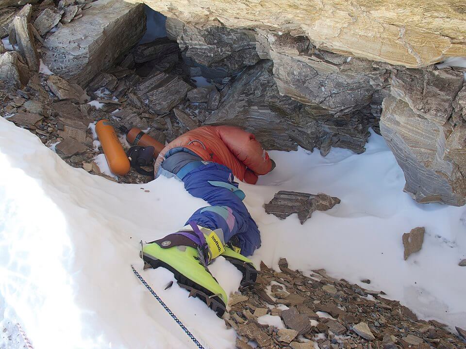 A deceased climber in colorful mountaineering gear lies partially covered in snow beneath a rocky overhang on a mountain slope, with oxygen tanks and equipment nearby.








