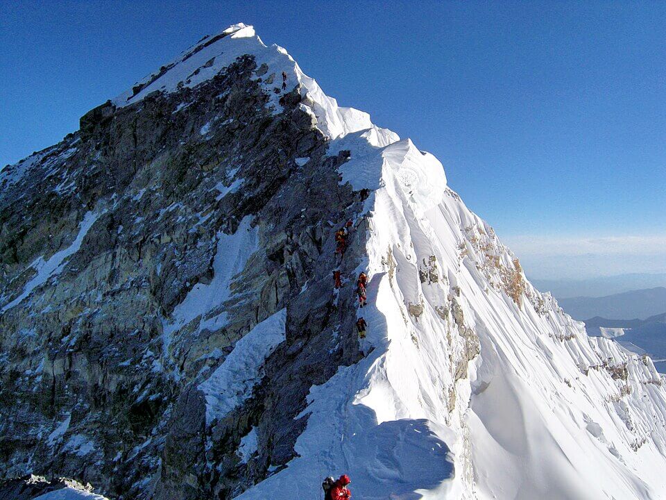 A line of climbers ascends a narrow, snow-covered ridge toward the summit of a steep, icy mountain peak under a clear blue sky, with deep drops on both sides.