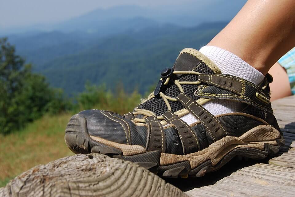 A close-up of a person’s foot wearing a rugged hiking sandal with white socks, resting on a wooden surface. In the background, forested hills and distant blue mountains fade into a hazy sky.