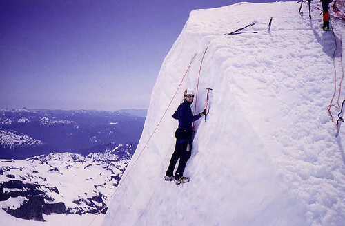 A climber in crampons and a helmet ascends a steep, snow-covered slope using an ice axe, secured by red ropes, with a panoramic view of snowy mountains and a clear sky in the background.
