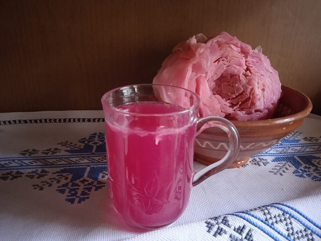 A clear glass mug filled with bright pink liquid sits on an embroidered cloth with a blue and black folk pattern. Behind it, a bowl holds a large, tightly packed pink fermented cabbage head.