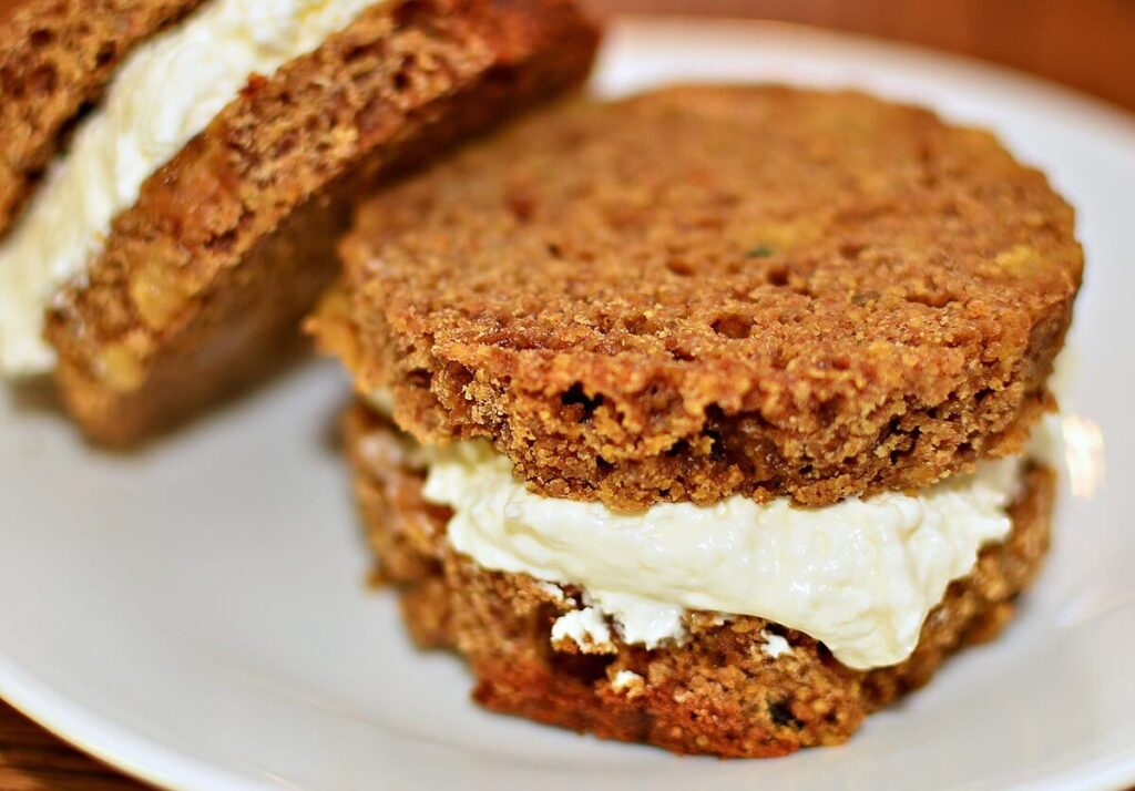 A close-up of a dessert sandwich made with two thick slices of carrot cake. The creamy white filling, likely cream cheese frosting, is generously spread between the moist, crumbly cake layers.