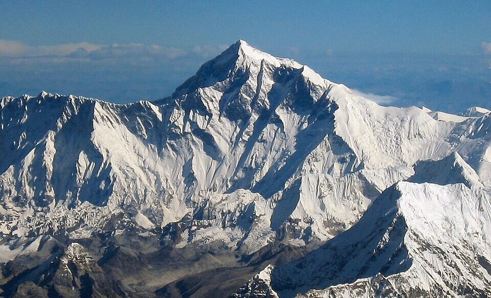 A clear, aerial view of Mount Everest, the world's tallest peak, towering above the surrounding snow-covered Himalayan mountain range under a blue sky.