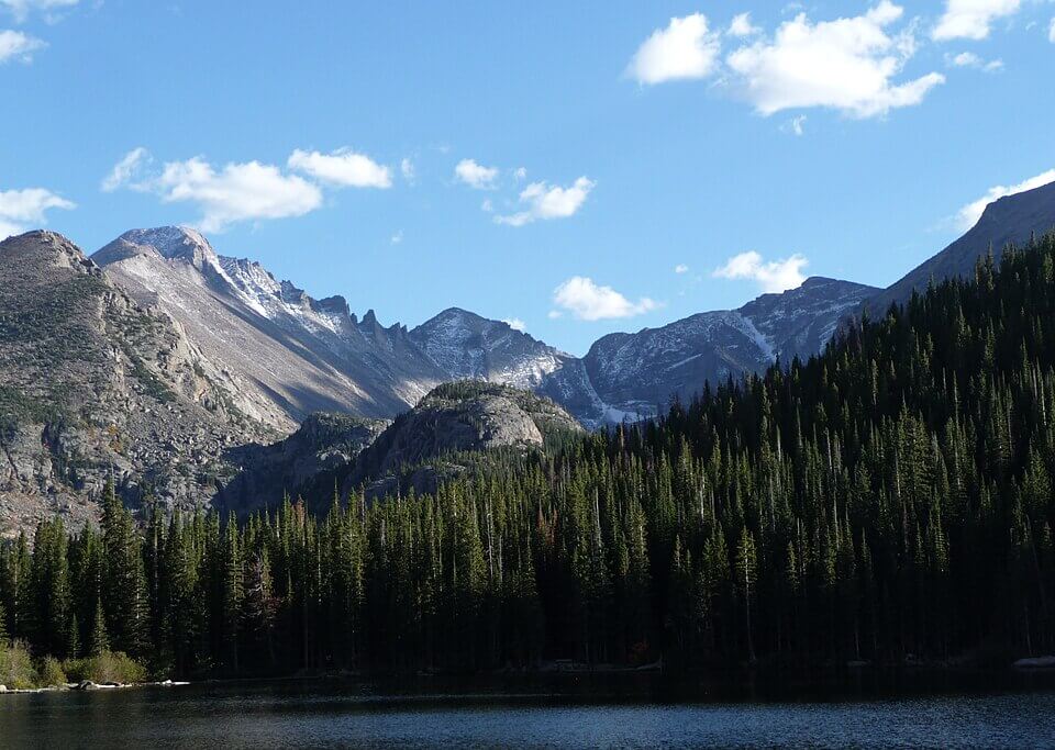 A scenic view of a mountain range with snow-capped peaks under a blue sky with scattered clouds. In the foreground, a dense forest of pine trees surrounds a calm, reflective lake.