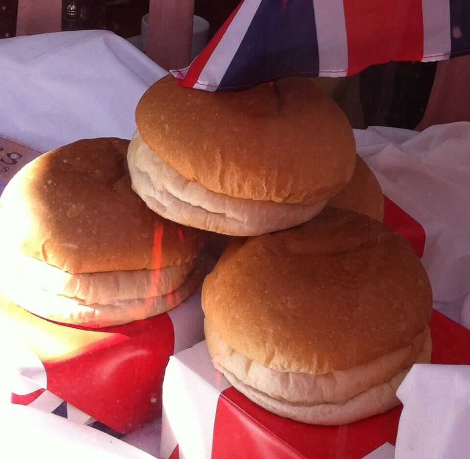 Four soft, round bread rolls stacked on red and white boxes with a British flag draped above them. The rolls have a light golden-brown top and a pale, fluffy bottom, displayed in a bright setting.