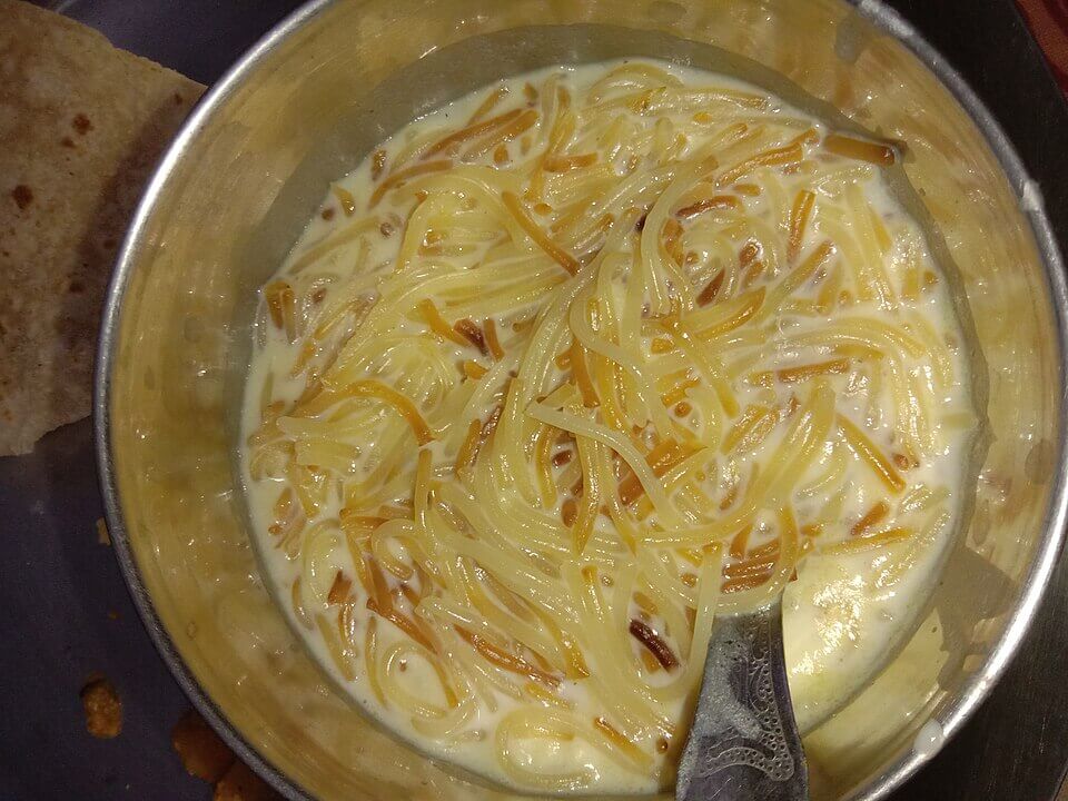 A metal bowl contains a creamy dessert made with thin vermicelli noodles soaked in milk. The mixture includes lightly toasted noodles, giving a mix of golden brown and pale yellow colors, with a spoon resting inside the bowl.