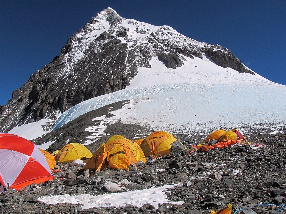 A cluster of brightly colored tents sits on a rocky, snowy slope beneath a towering ice-covered mountain peak, with a glacier visible in the middle ground under a deep blue sky.







