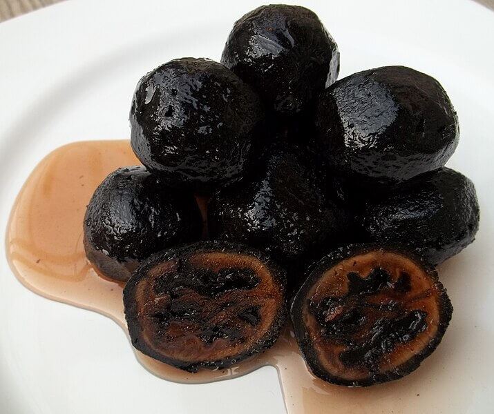 A pile of glossy, black preserved walnuts on a white plate, with syrup pooling underneath. Two of the walnuts are cut open, revealing their dark, textured interiors.